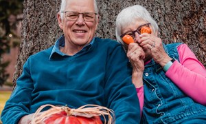 elderly couple leaning on a tree trunk holding colourful pumpkins