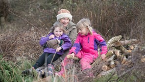 smiling grandparent with two young children sitting on a grassy bank next to a pile of logs